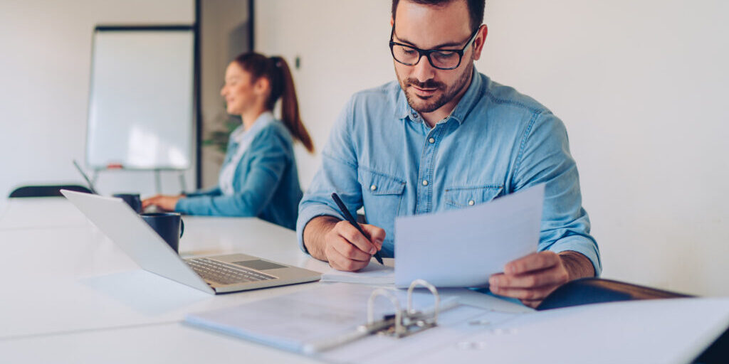 Young businessman wearing denim shirt and eyeglasses doing paperwork while sitting at the desk in the big white office with coworkers