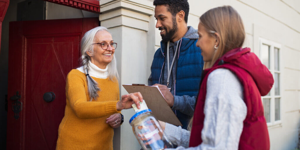 Young door to door fundraisers talking to senior woman and collecting money for charity in a street.